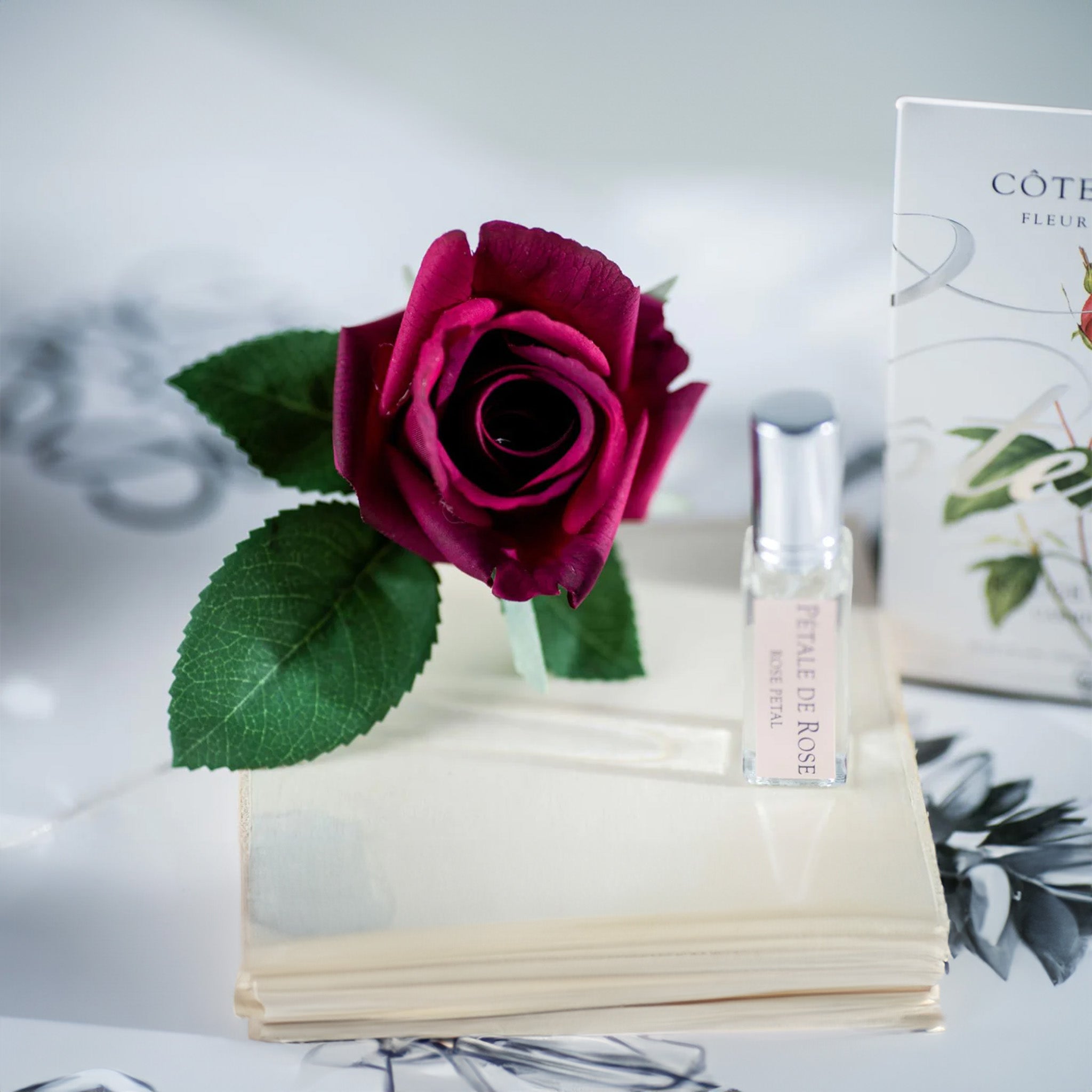 A carmine red rosebud on an open book with a clear perfume vial and a white 'CÔTE NOIRE' box in a soft-focus background.