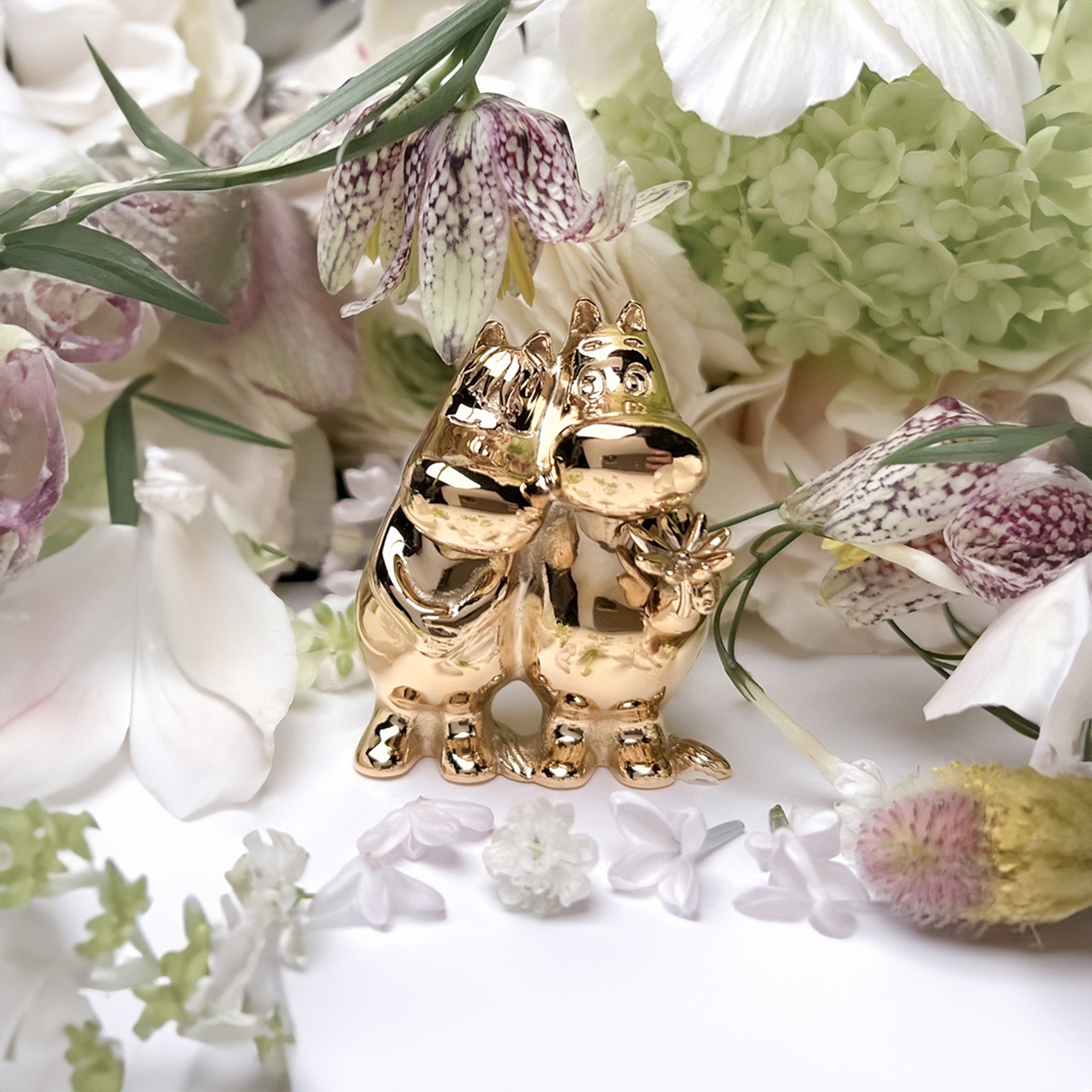 a gold Moomintroll and his beloved Snorkmaiden hugging on top of a table surrounded by flowers