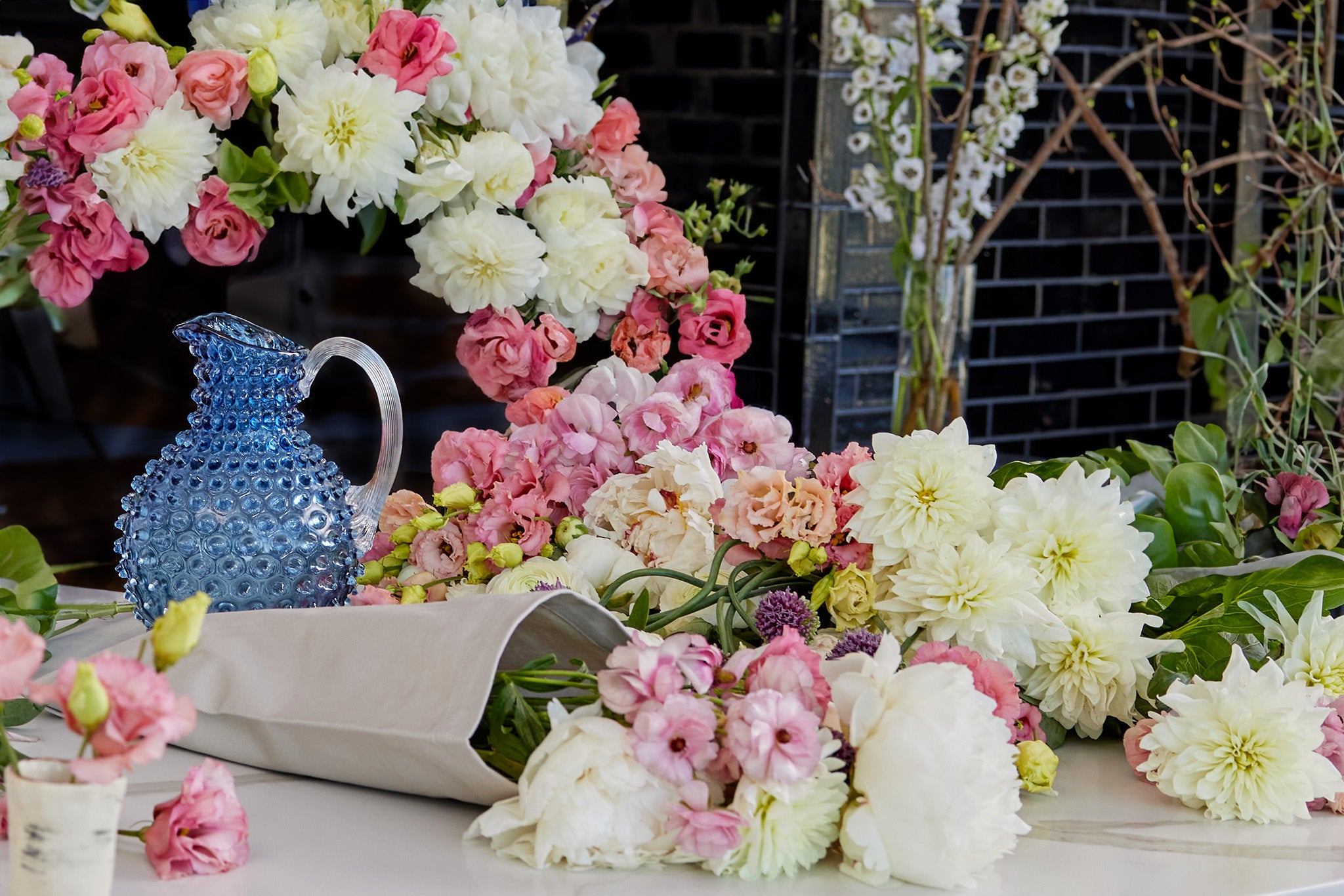 A vibrant and colorful floral arrangement featuring an array of pink, white, and peach flowers, with a distinctive blue textured glass jug in the center, set against a backdrop with soft focus greenery and a dark lattice structure.