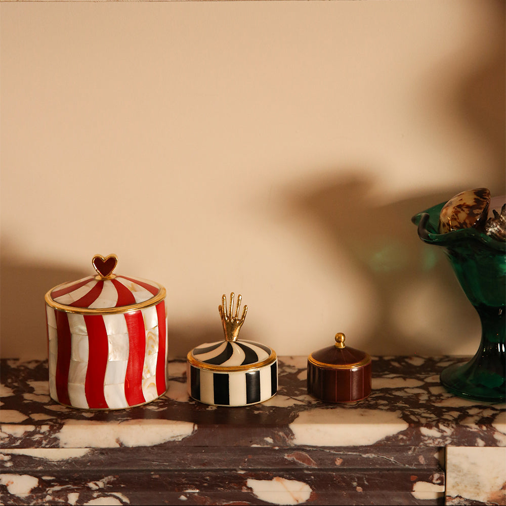 Three decorative lidded jars with circus-inspired patterns are arranged on a marble surface against a beige wall. The left jar has red and white vertical stripes with a gold heart lid; the center jar has black and white stripes with a gold crown lid; the right jar is dark brown with a gold lid.
