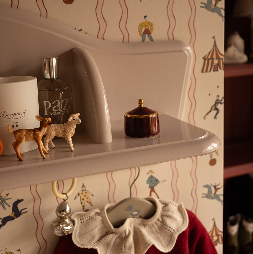 A close-up of a beige molded shelf or tray holding a white mug with 'Beepboots' text, a dark bottle labeled 'Paz de Luxe', two small deer figurines, and a dark round box with a gold lid. The background is patterned wallpaper with whimsical figures and wavy lines, and a red garment with a cream ruffled collar hangs below.