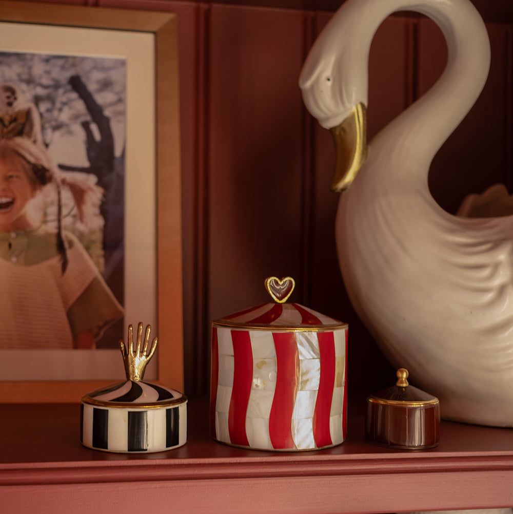 A decorative red and white striped lidded box with a gold heart accent on top, styled on a reddish-brown shelf alongside other ornate home decor items including a white swan sculpture and a framed print.