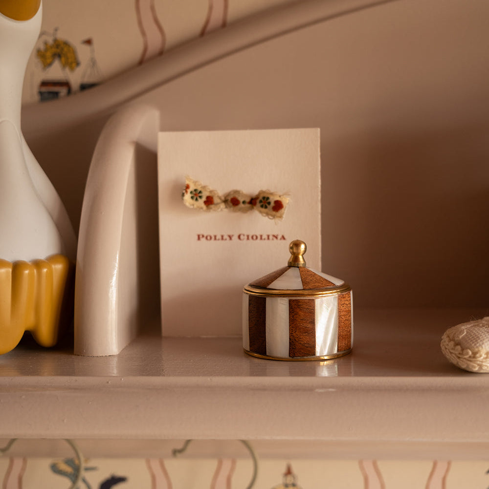 A close-up of a jewelry display card featuring a small gold-toned accessory with colorful floral details, alongside a small striped lidded container on a shelf in a warm beige setting.