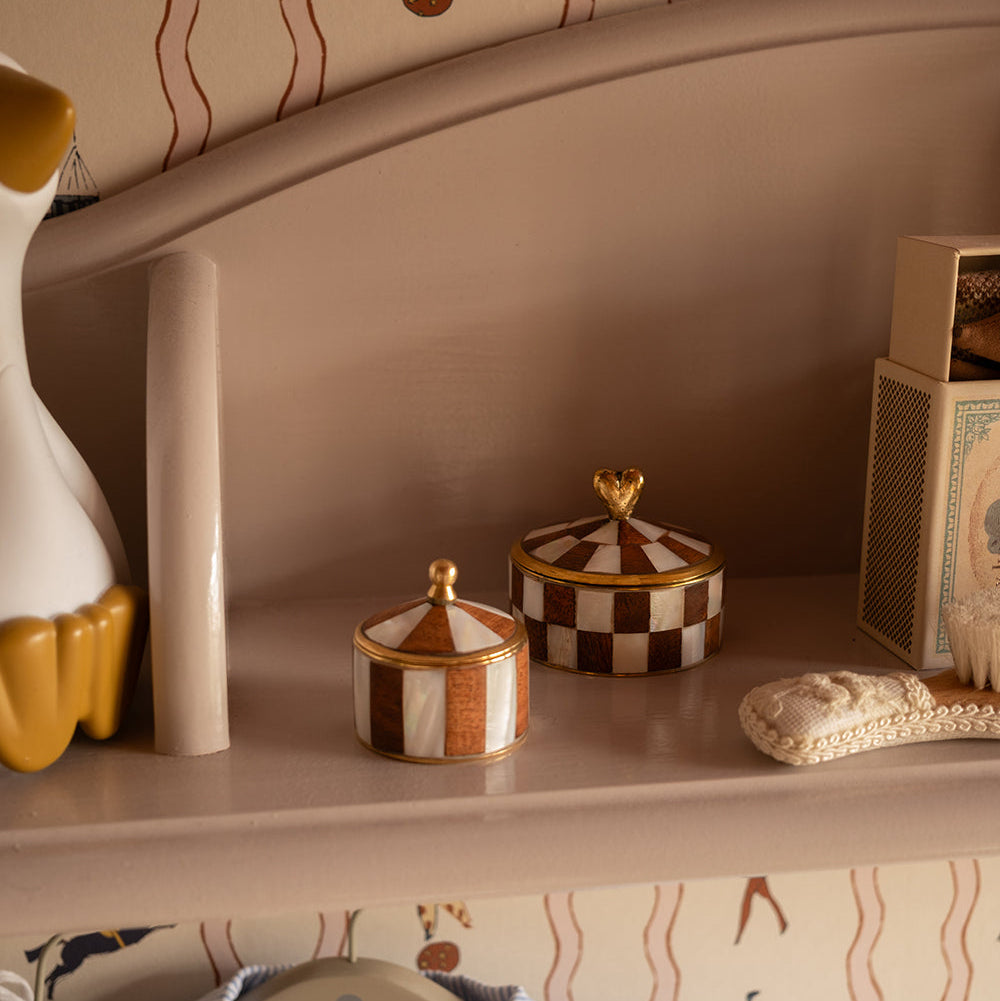 Two small decorative lidded boxes with a brown-and-white checkered pattern and gold trim sit on a beige shelf, surrounded by nursery-style decor and patterned wallpaper.