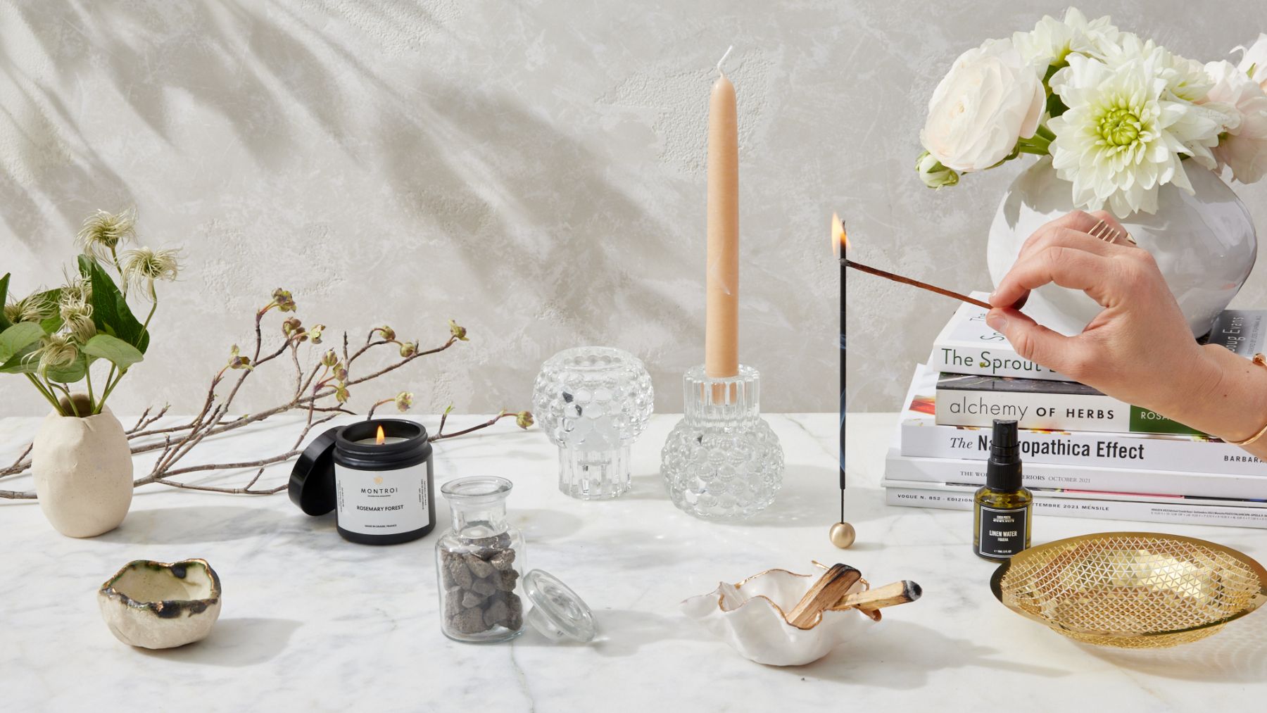 This image features a serene tabletop scene with a hand lighting an incense stick over a golden tray. Nearby, there’s a stack of books, including “The Sprout Book.” The table is adorned with a beige candle in a hobnail glass candle holder, a black “Rosemary Forest” candle by Montroi, a bottle of linen water from Casa Posta, a vase with fresh flowers, branches, and a small jar of stones, creating a calming and natural ambiance.