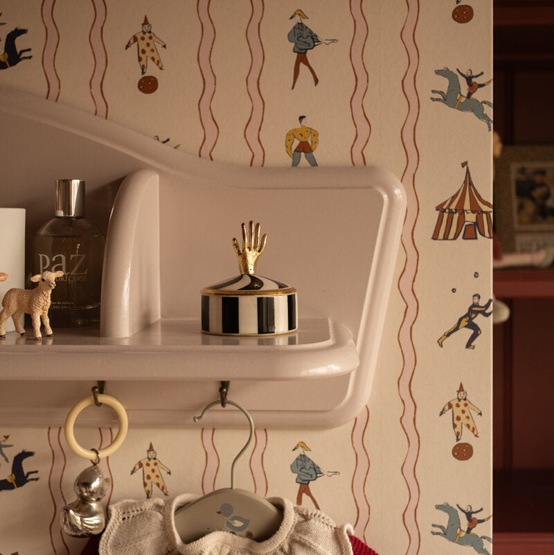 A styled interior scene showing a cream-colored shelf against wallpaper with a circus-themed print. The wallpaper features red wavy lines and illustrations of performers, horses, and a tent.