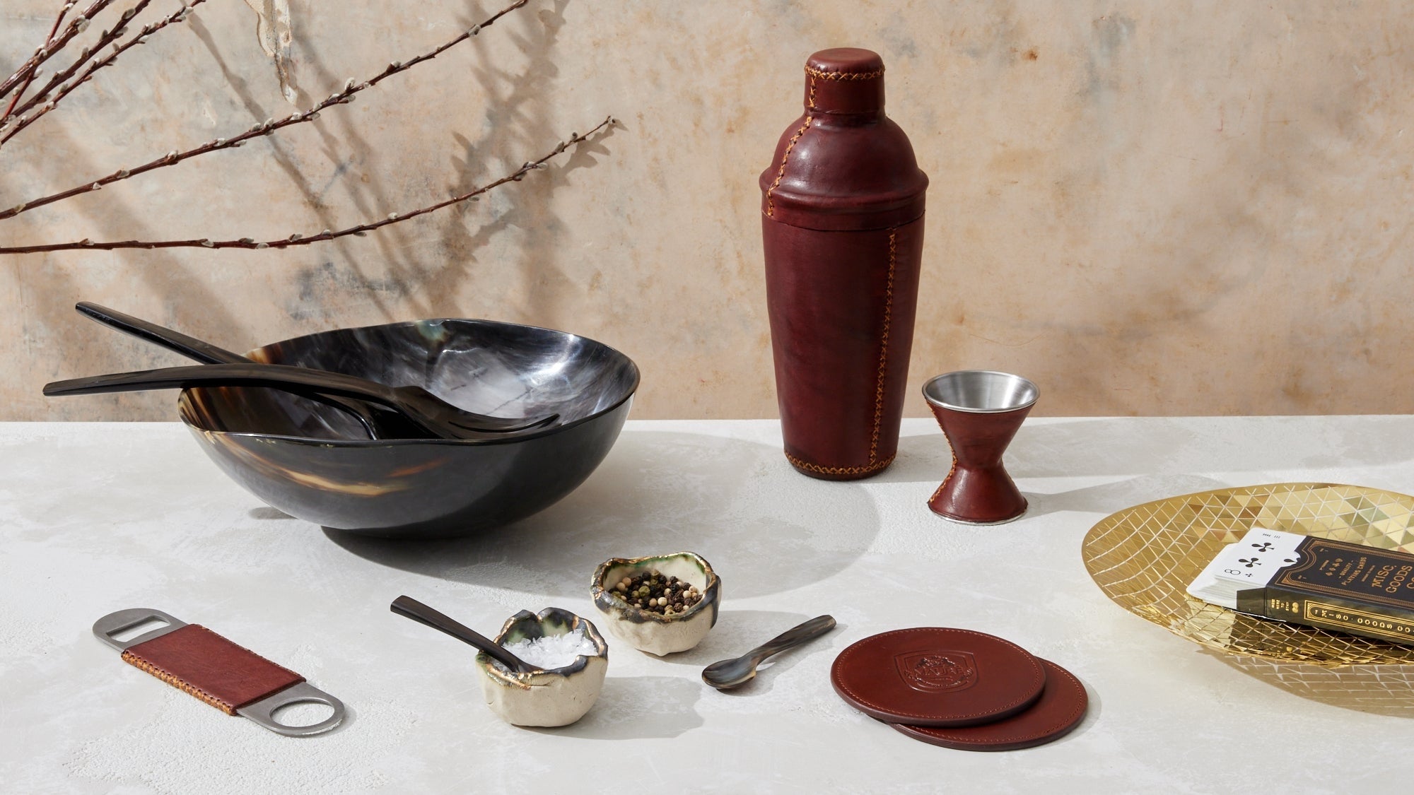 This image showcases an elegant collection of tabletop accessories. A horn bowl with matching serving utensils takes center stage, accompanied by small ceramic dishes filled with salt and pepper. Nearby, a leather-wrapped cocktail shaker and matching jigger, along with leather coasters embossed with the Maure logo, add a touch of sophistication. A gold-toned tray by Fundamental Berlin holds a deck of cards, completing the stylish and luxurious setting. The scene blends natural materials with refined craftsm