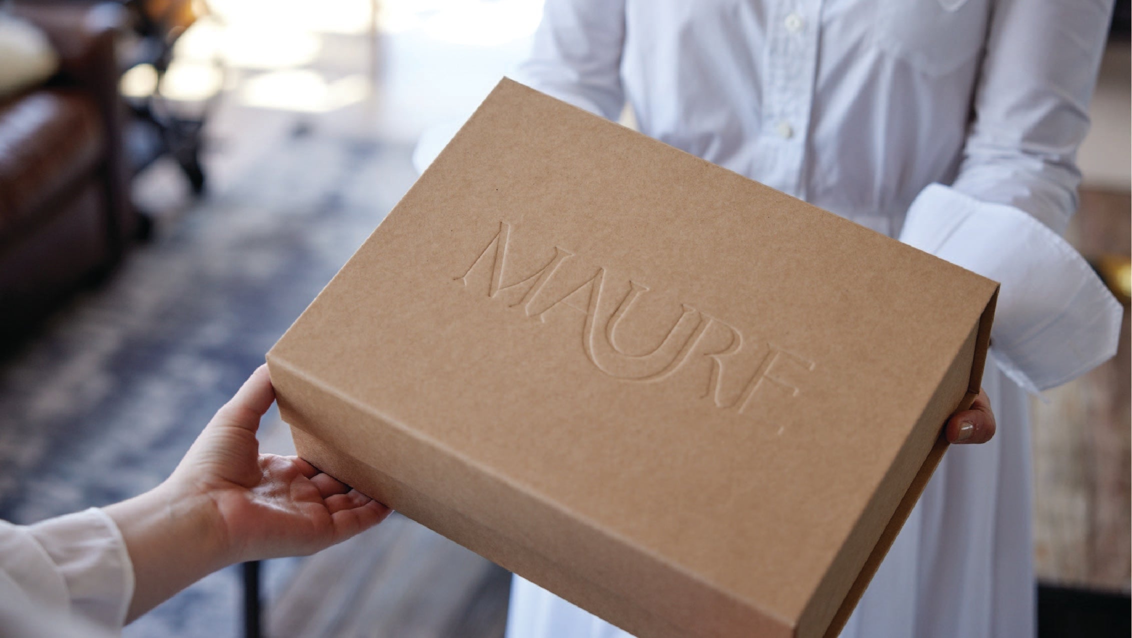 This image shows a person handing over a brown gift box embossed with the “Maure” logo. The scene suggests a moment of gifting or receiving, with the box being held between two people dressed in white. The background is softly blurred, focusing attention on the elegantly designed box, which conveys a sense of luxury and thoughtful presentation.