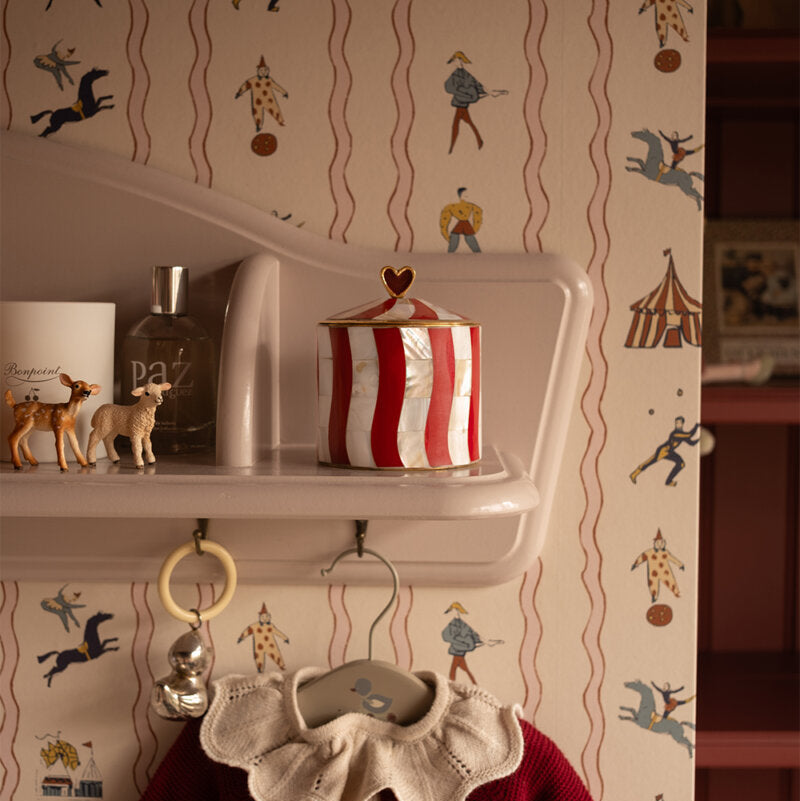 A styled interior scene showing a red and white striped tin with a gold heart lid on a white shelf, with circus-themed wallpaper in the background and a burgundy sweater hanging below.