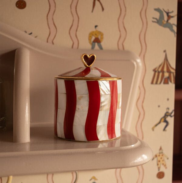 A small cylindrical lidded box with red and white vertical stripes and gold trim sits on a cream-colored shelf. The lid has a small gold heart finial.