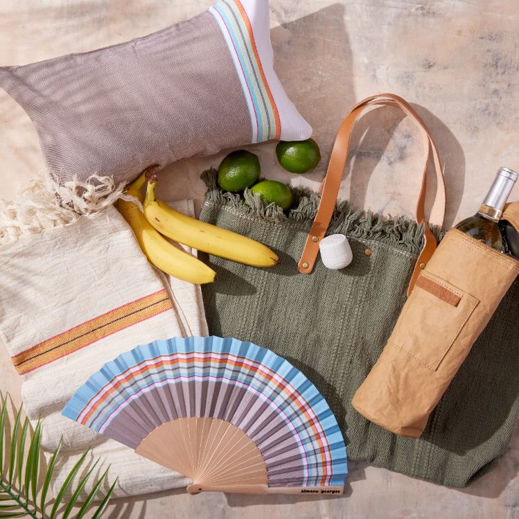 This image features a laid-back, resort vibe with a collection of items arranged on a light surface. There’s a woven pillow with colorful stripes, a pair of bananas, limes, and an olive green tote bag with leather straps. The bag holds a small white container, and next to it is a bottle of wine in a tan wine carrier. A folded beach towel with a red and yellow stripe and a blue-striped hand fan complete the luxurious, relaxed setting.