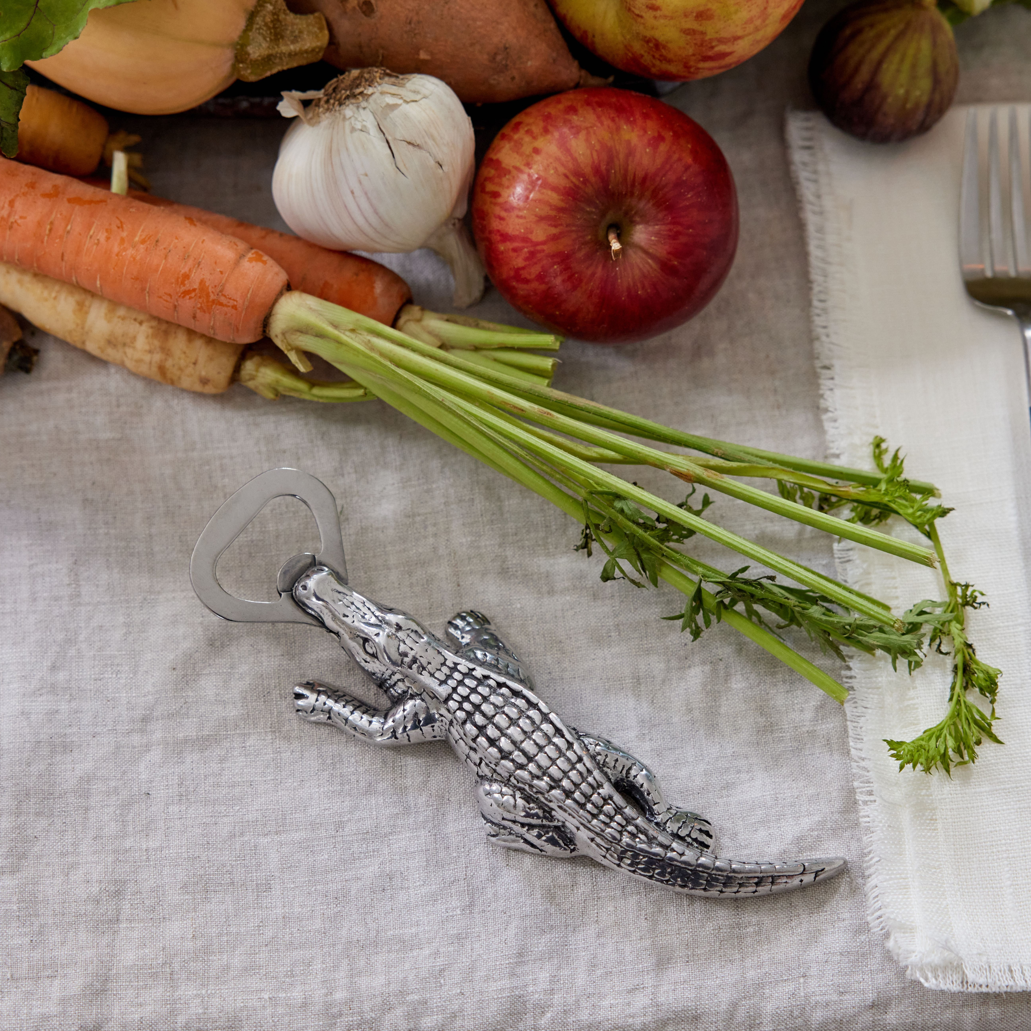 a pewter alligator bottle opener on a linen table surrounded by fruit and vegetables 