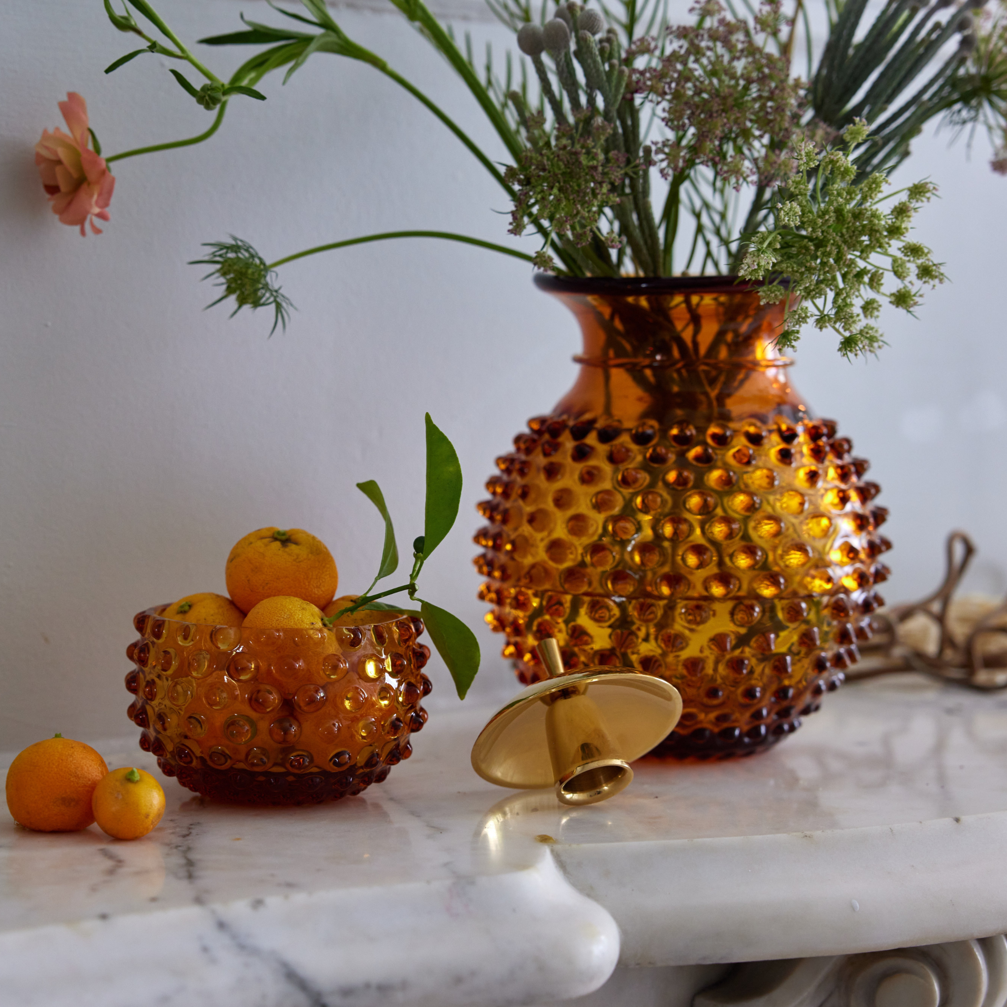 a marble table topped with an amber hobnail bowl filled with oranges and an amber hobnail vase filled with flowers