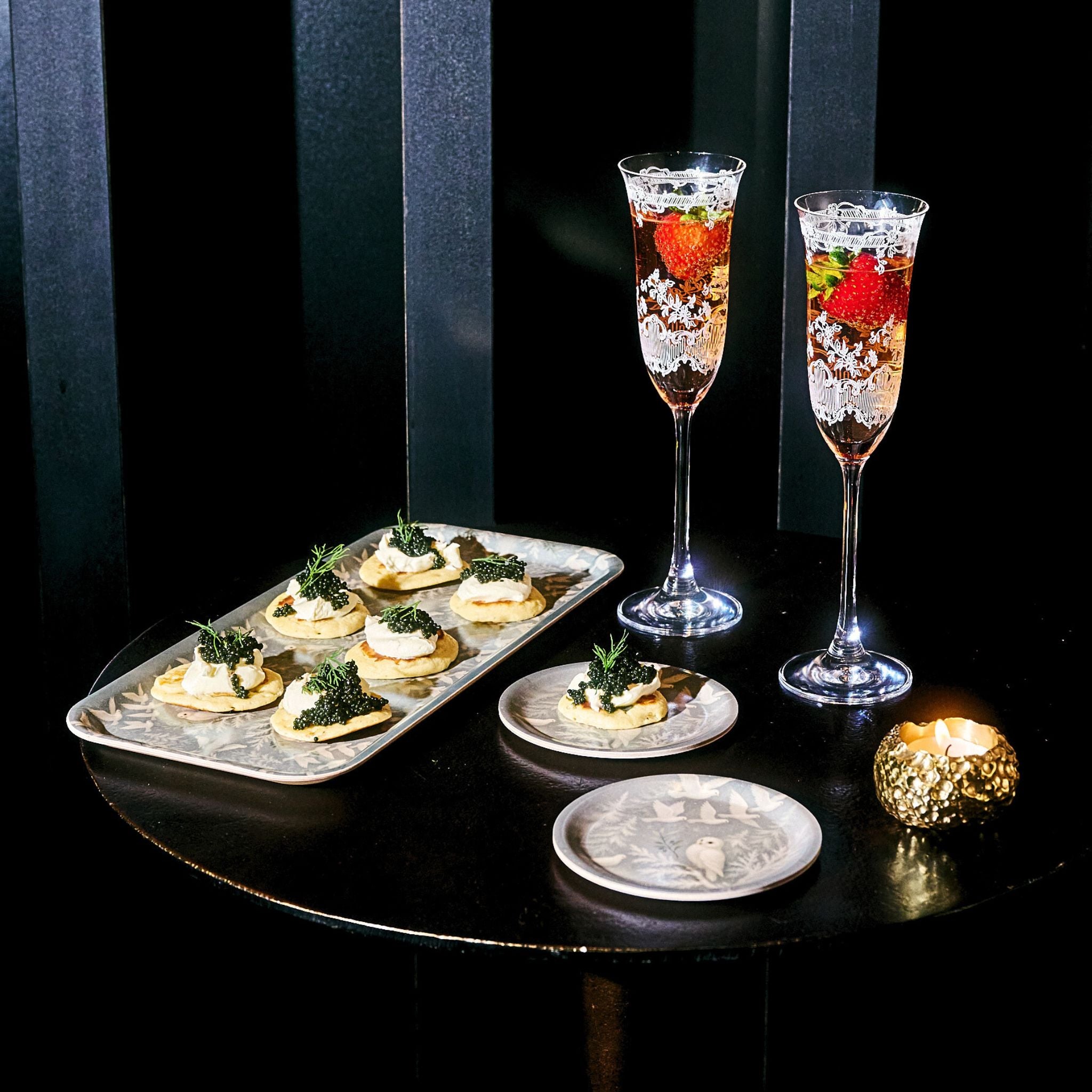 Two champagne flutes with garnishes on a black table, accompanied by small appetizers.