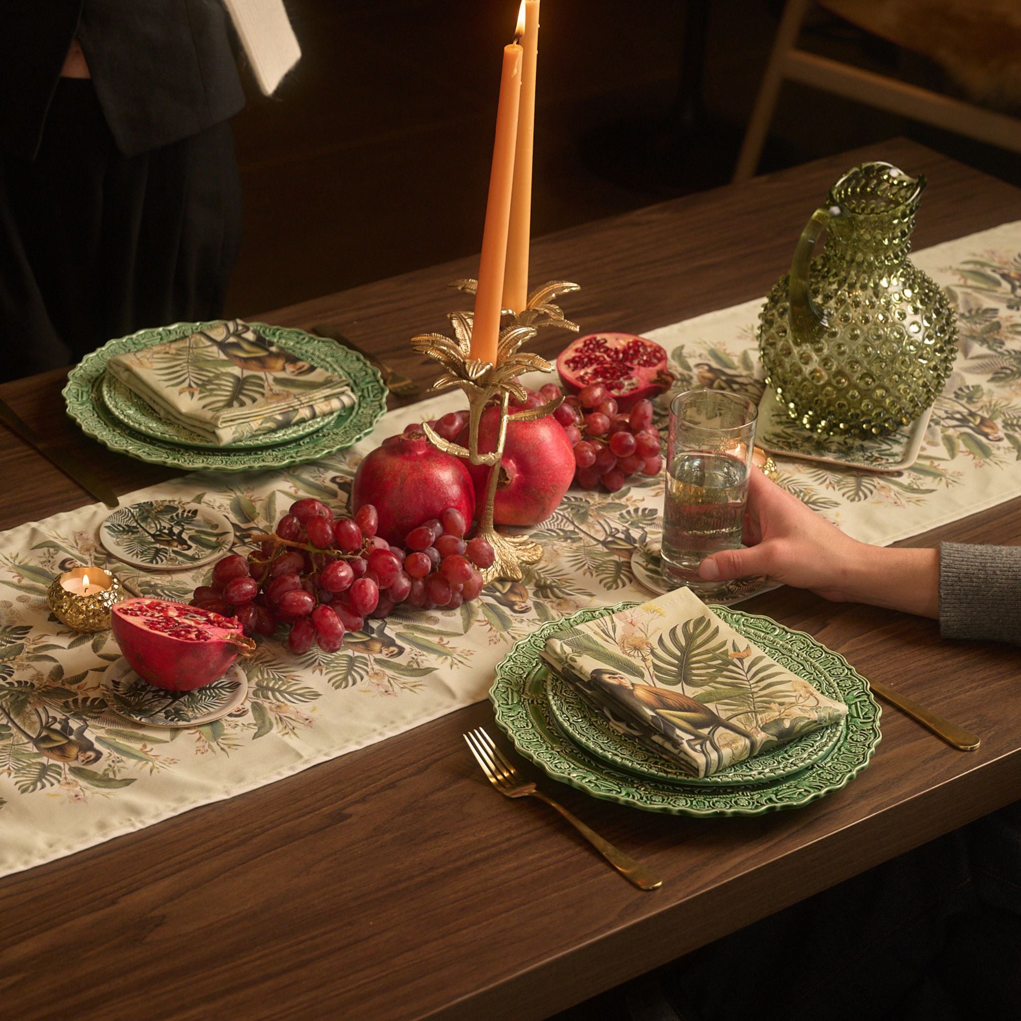 Dinner table setting with green plates, pomegranates, grapes, and a candle on a decorative tablecloth.