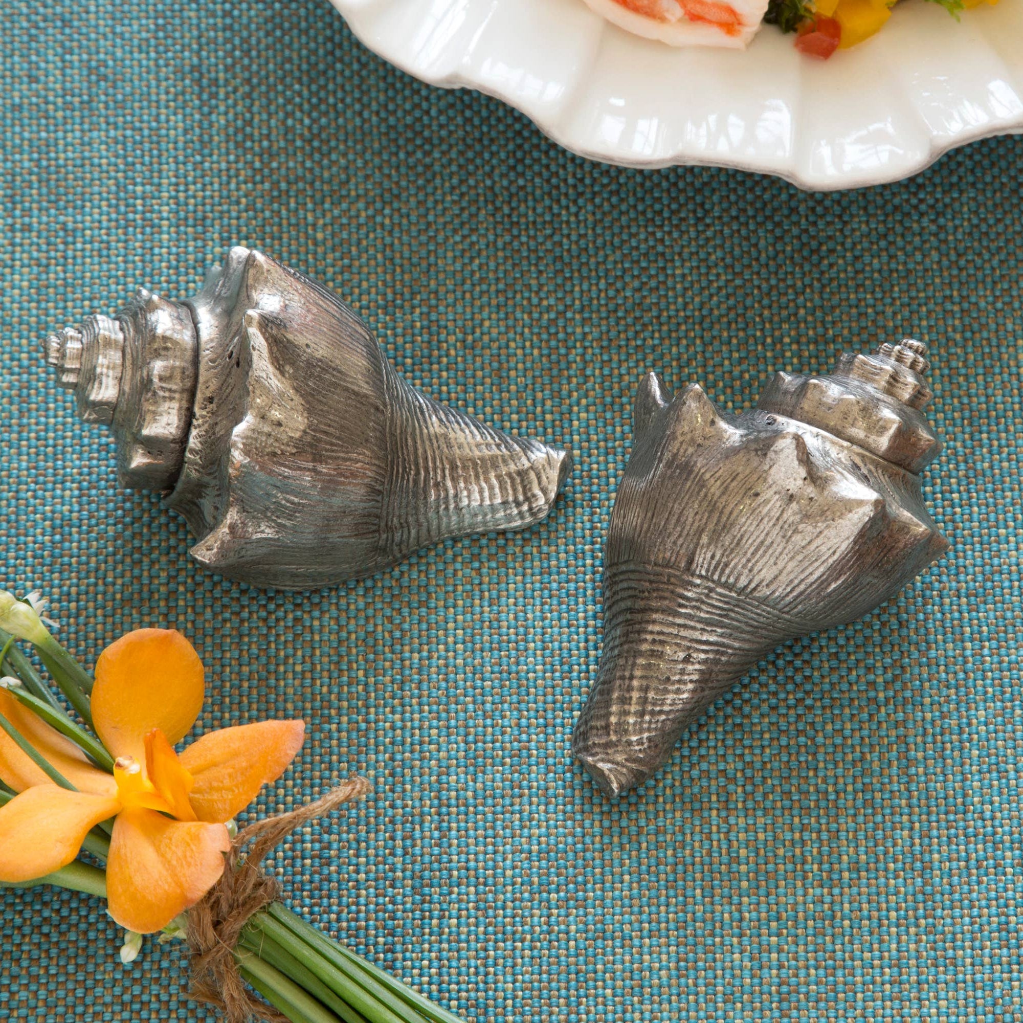 Two metallic shells on a textured blue surface with flowers and a decorative plate in the background.