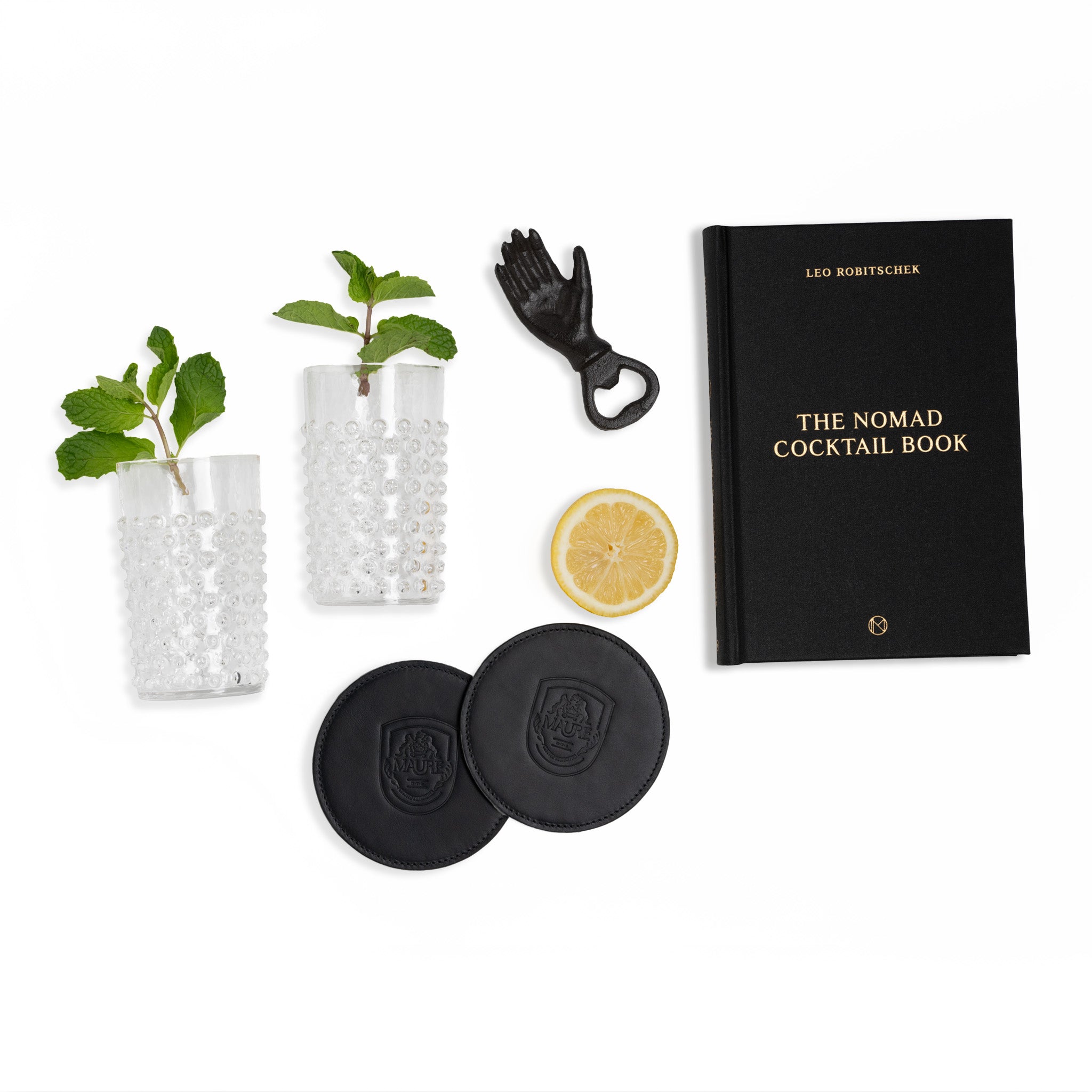 This image showcases a stylish and sophisticated gift collection perfect for cocktail enthusiasts. The set includes “The Nomad Cocktail Book” by Leo Robitschek, two clear hobnail glasses, a unique hand-shaped bottle opener, and two black leather coasters embossed with the Maure logo. A slice of lemon and fresh mint sprigs are also included, adding a fresh and vibrant touch to the ensemble. This collection is ideal for someone who enjoys mixing drinks and appreciates high-quality barware and accessories.