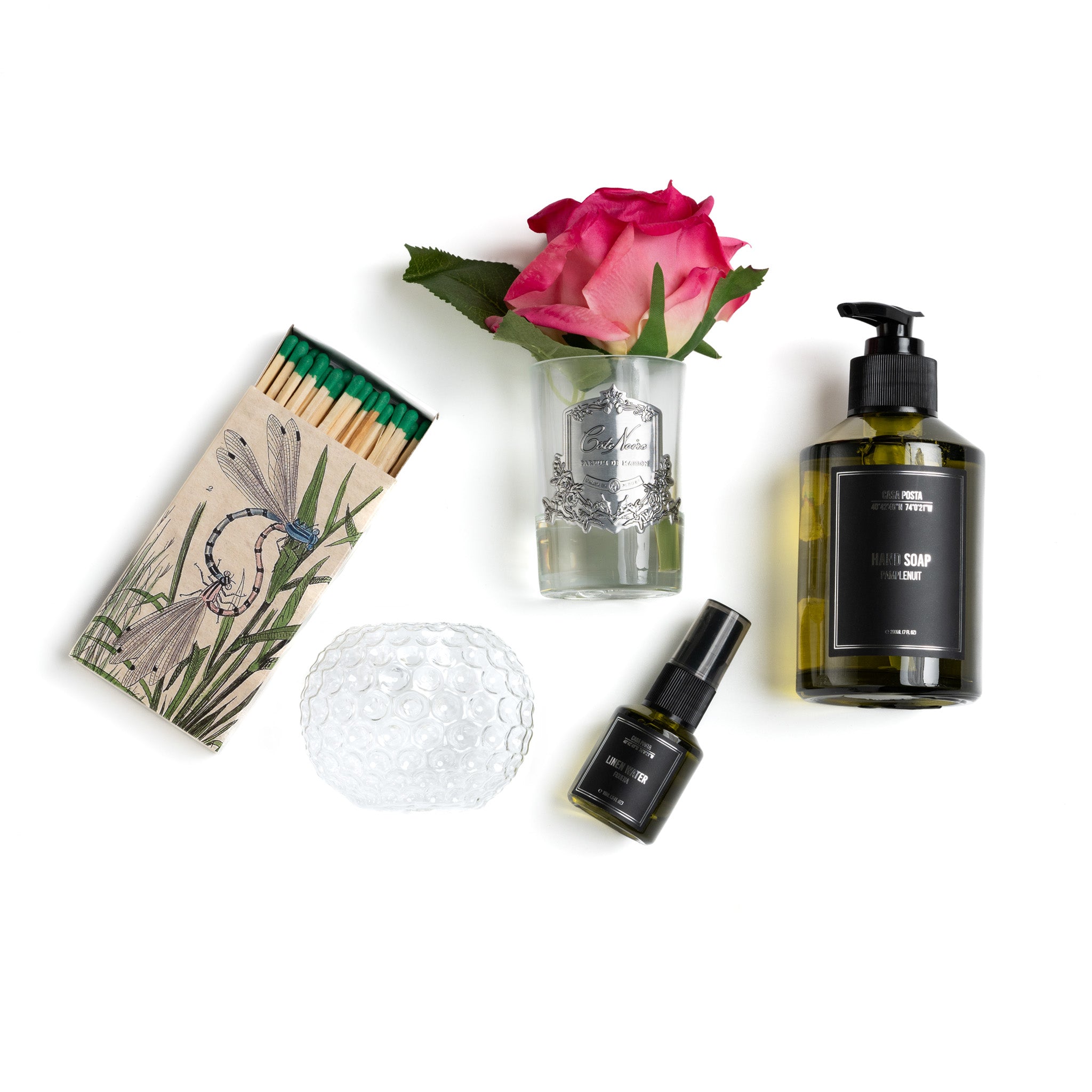 This image showcases a beautifully curated collection of scented and decorative items. The set includes a box of matches with a nature-themed design, a bubble glass candle holder, a bottle of hand soap by Casa Posta, a small spray bottle of linen water by Casa Posta, and a pink rose arranged in a delicate glass vase by Cote Noire. This collection exudes elegance and sophistication, making it perfect for someone who appreciates refined scents and luxurious home decor.