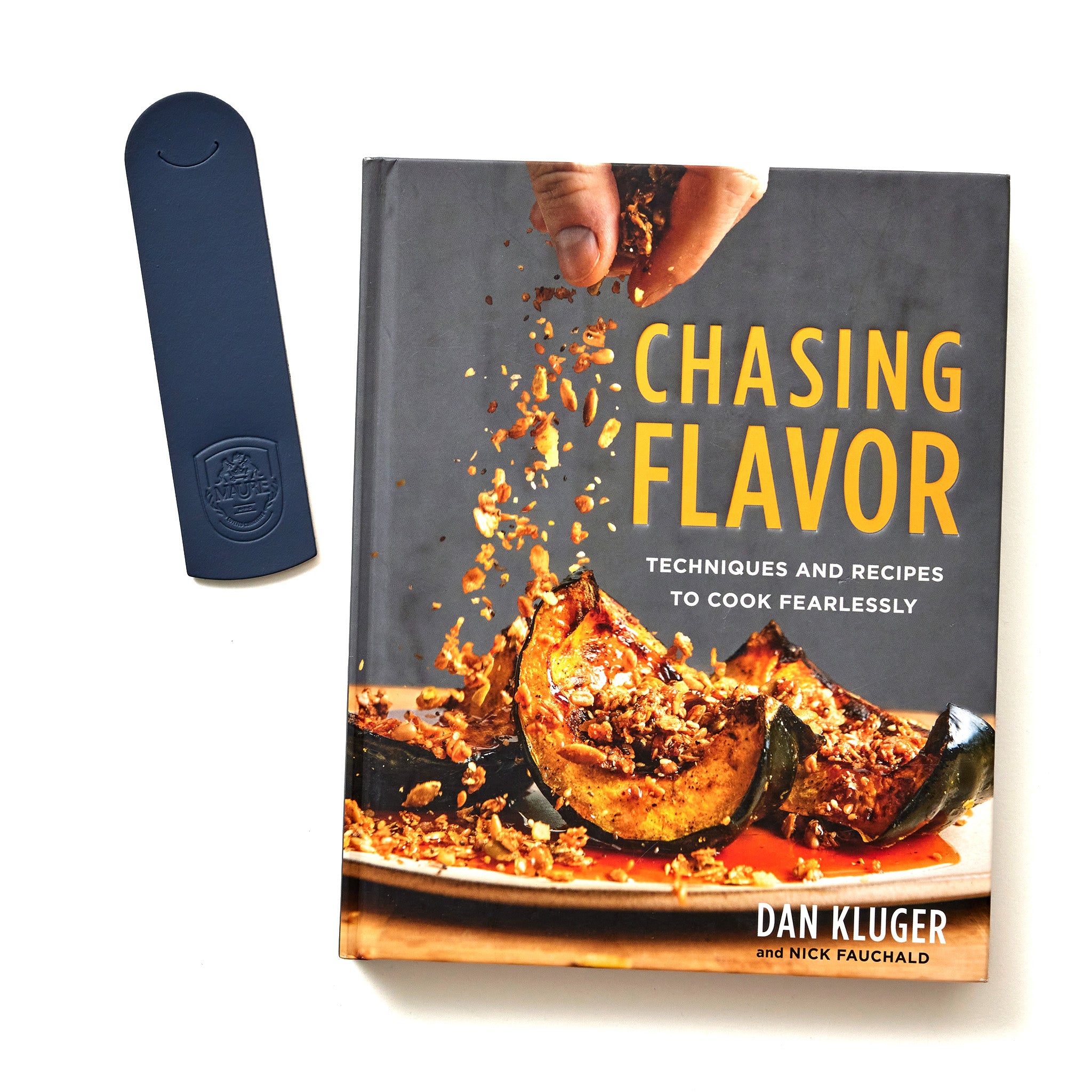 A cookbook titled 'Chasing Flavor: Techniques and Recipes to Cook Fearlessly' by Dan Kluger and Nick Fauchald, with a navy blue bookmark placed beside it. The cover features an image of hands sprinkling toppings onto roasted squash. The bookmark has the Maure crest embossed on it. The book and bookmark are set against a white background, highlighting their details.