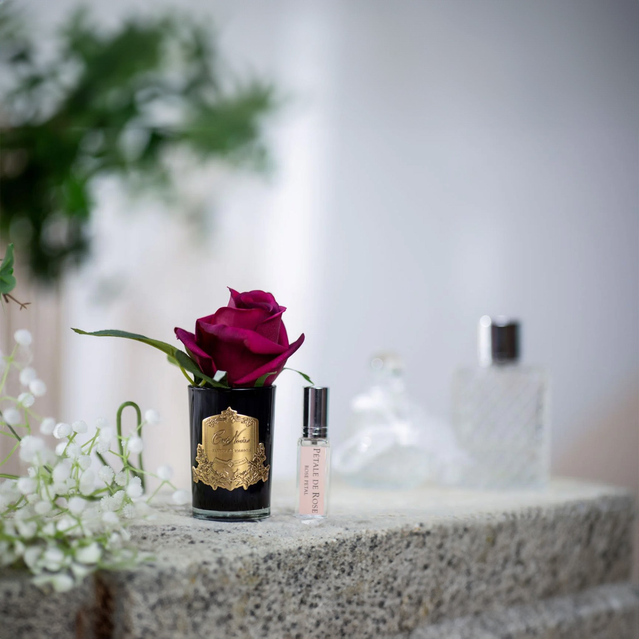 A carmine red rosebud in a black vase with a gold label on a stone surface with white flowers and a small perfume vial in the background