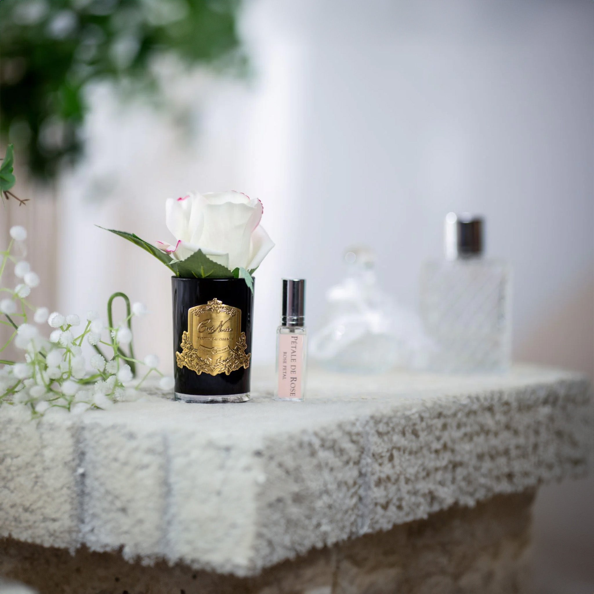 a stone table with a black vase of a blush pink rose and a bottle of fragrance by cote noire