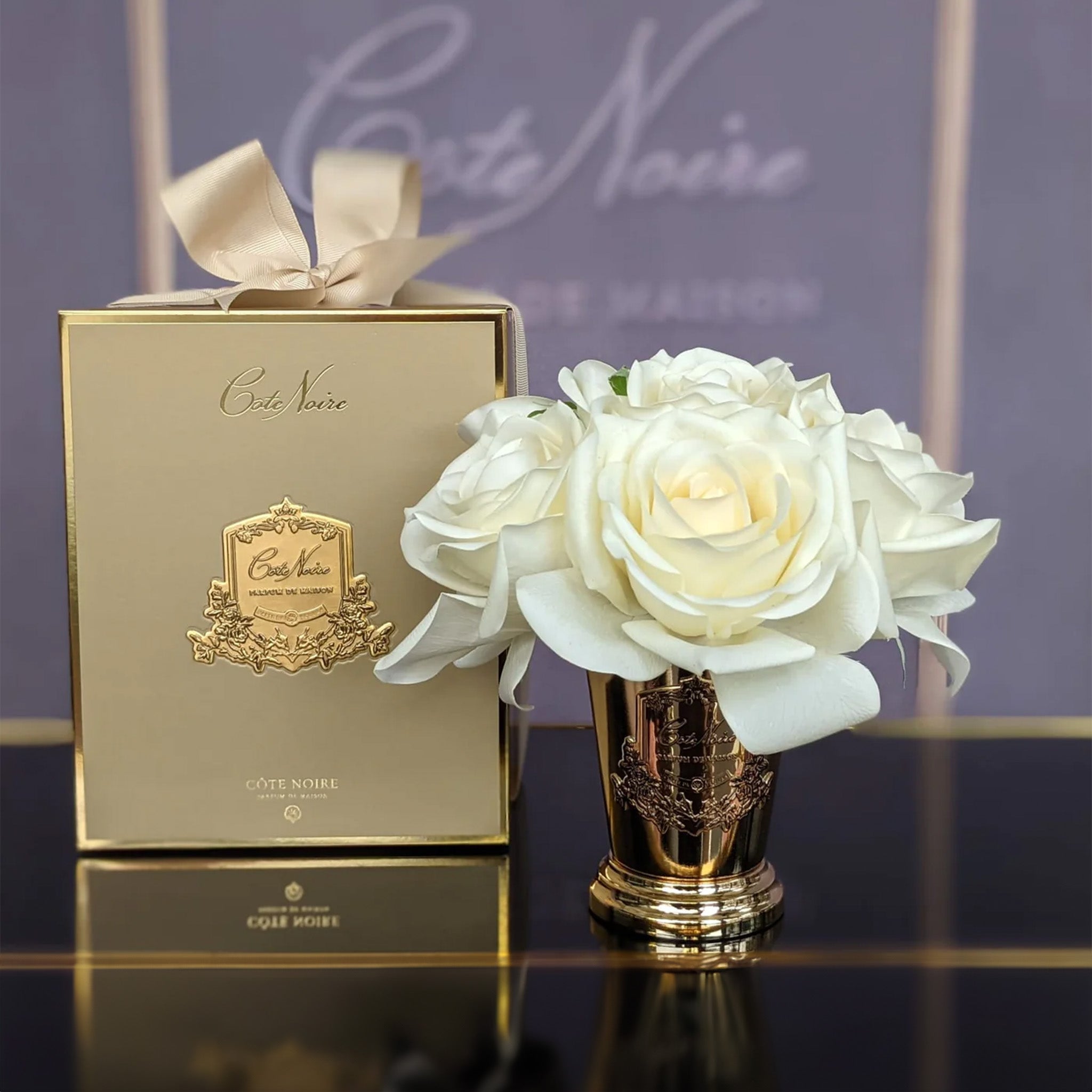 a white rose bouquet in a gold vase next to a gold box by cote noire