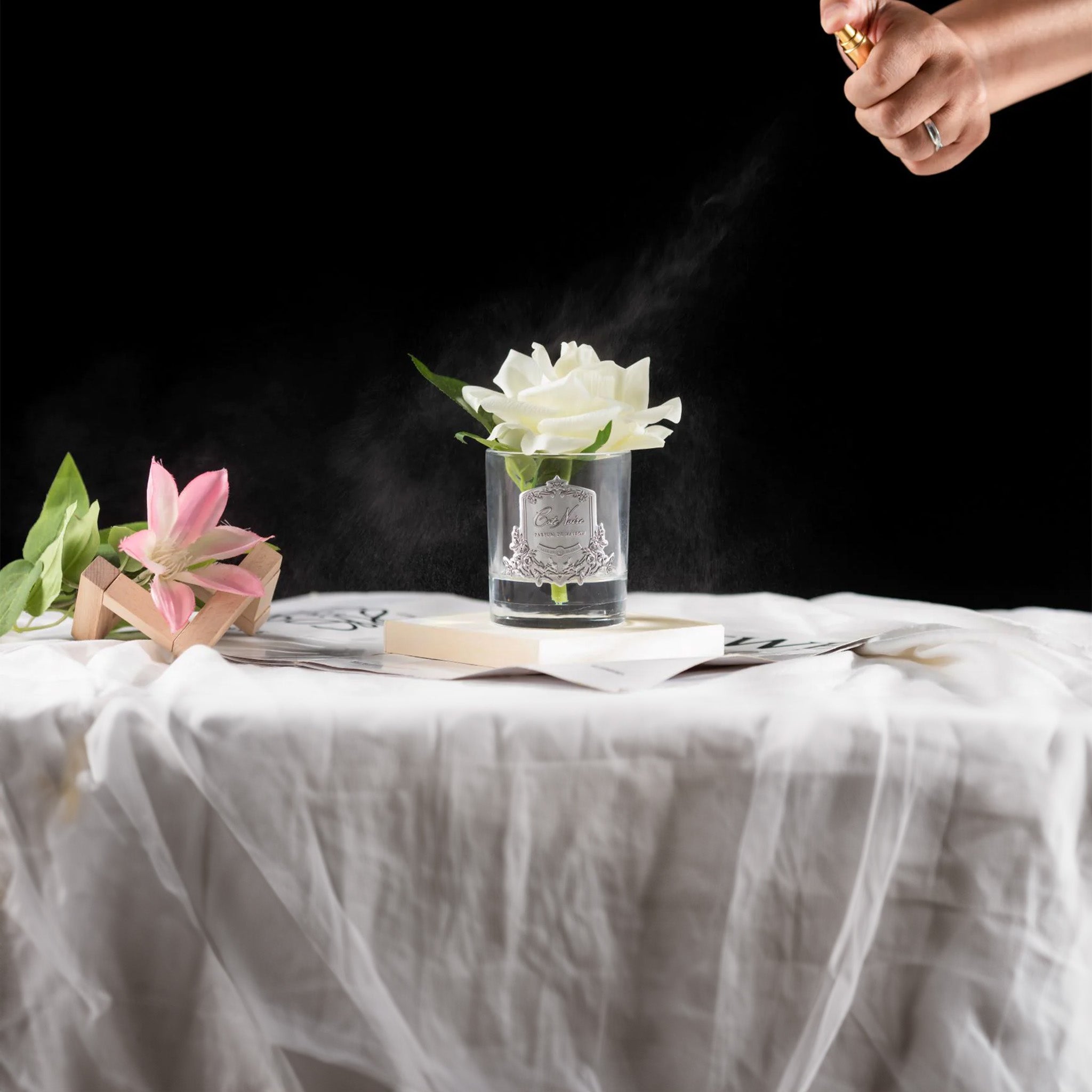 a table topped with a person spritzing fragrance on a white rose in a glass vase by cote noire