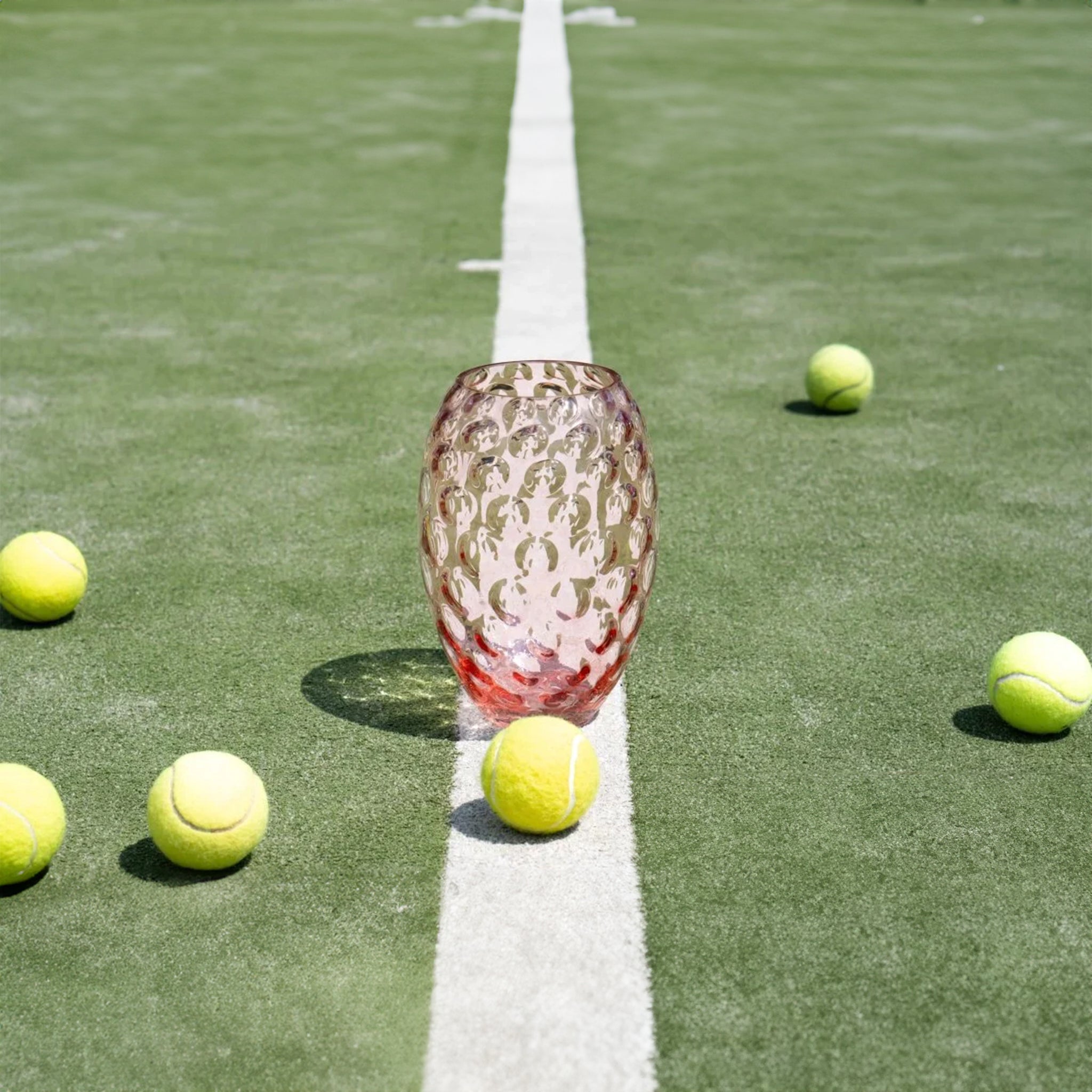 a row of tennis balls on a tennis court surrounding a pink vase 