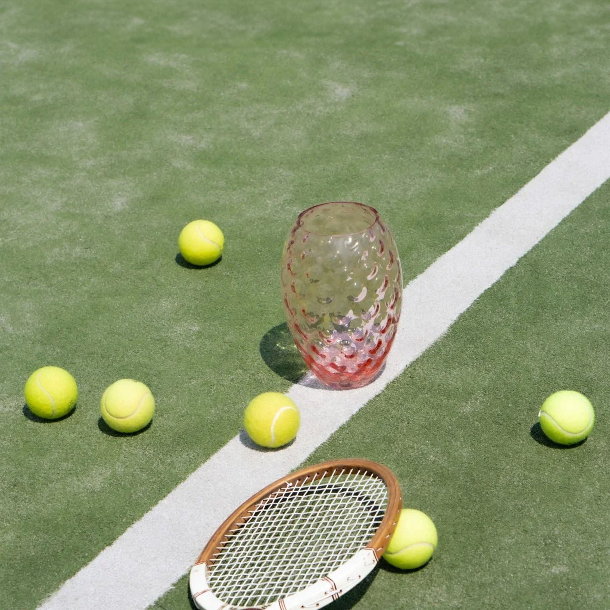 a tennis racket, balls, and pink vase on a tennis court