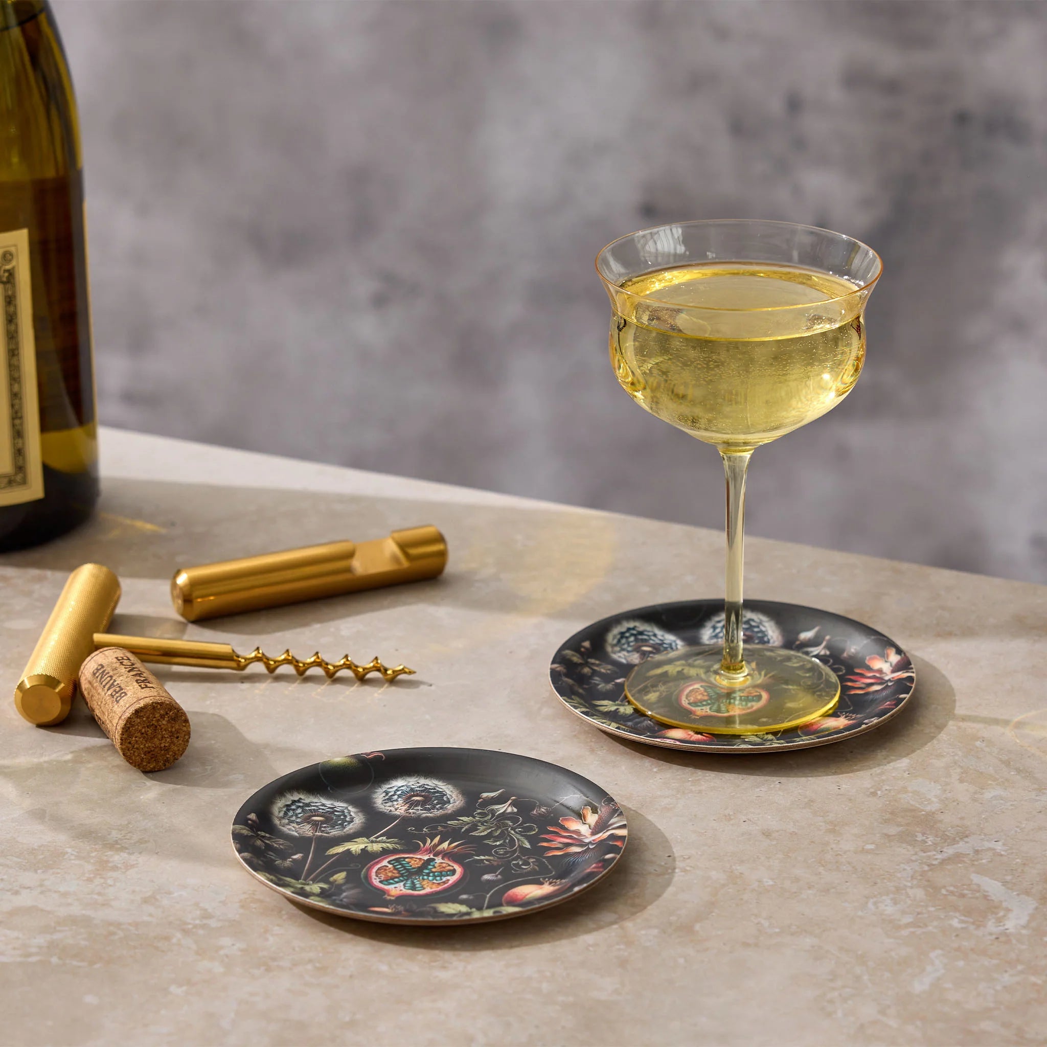 Wine glass with a floral coaster on a stone surface, accompanied by a bottle, cork, and corkscrew.
