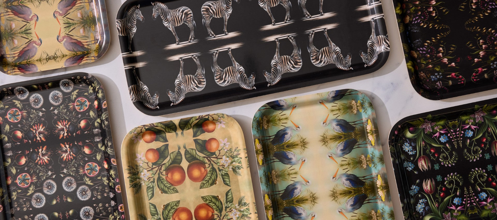 This image features a collection of birchwood trays by Maure, each with a unique design. The trays display a variety of patterns, including zebras on a black background, oranges with green leaves on a cream background, exotic birds against a nature scene, and intricate floral and fruit patterns on darker backgrounds. The trays are arranged on a light surface, showcasing their vibrant and diverse designs.