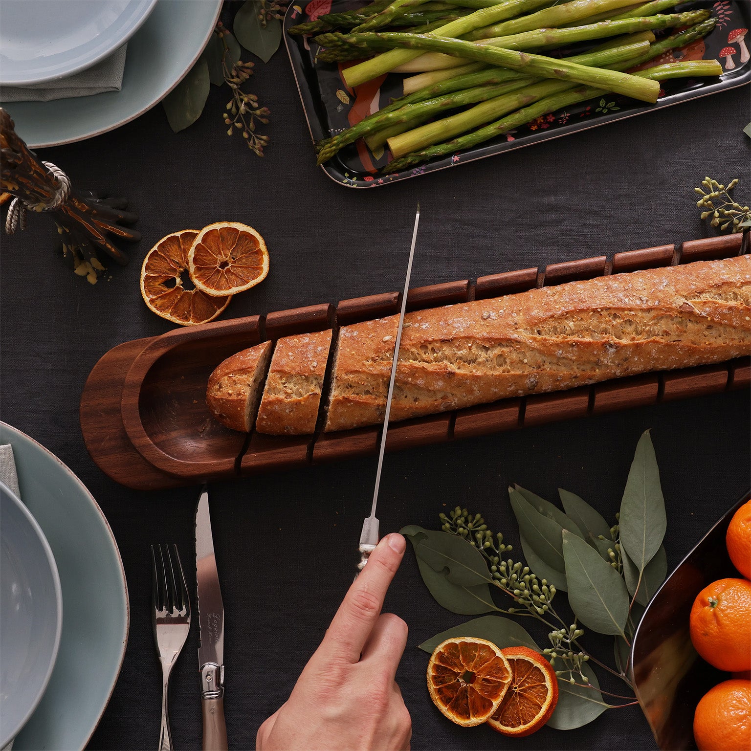 This image captures a cozy dining scene with a hand slicing a baguette on a wooden breadboard. The table is set with plates, silverware, and a side dish of roasted asparagus on a decorative tray. Slices of dried oranges and fresh mandarins in a metal bowl add a touch of color, while eucalyptus leaves contribute to the natural, rustic ambiance.