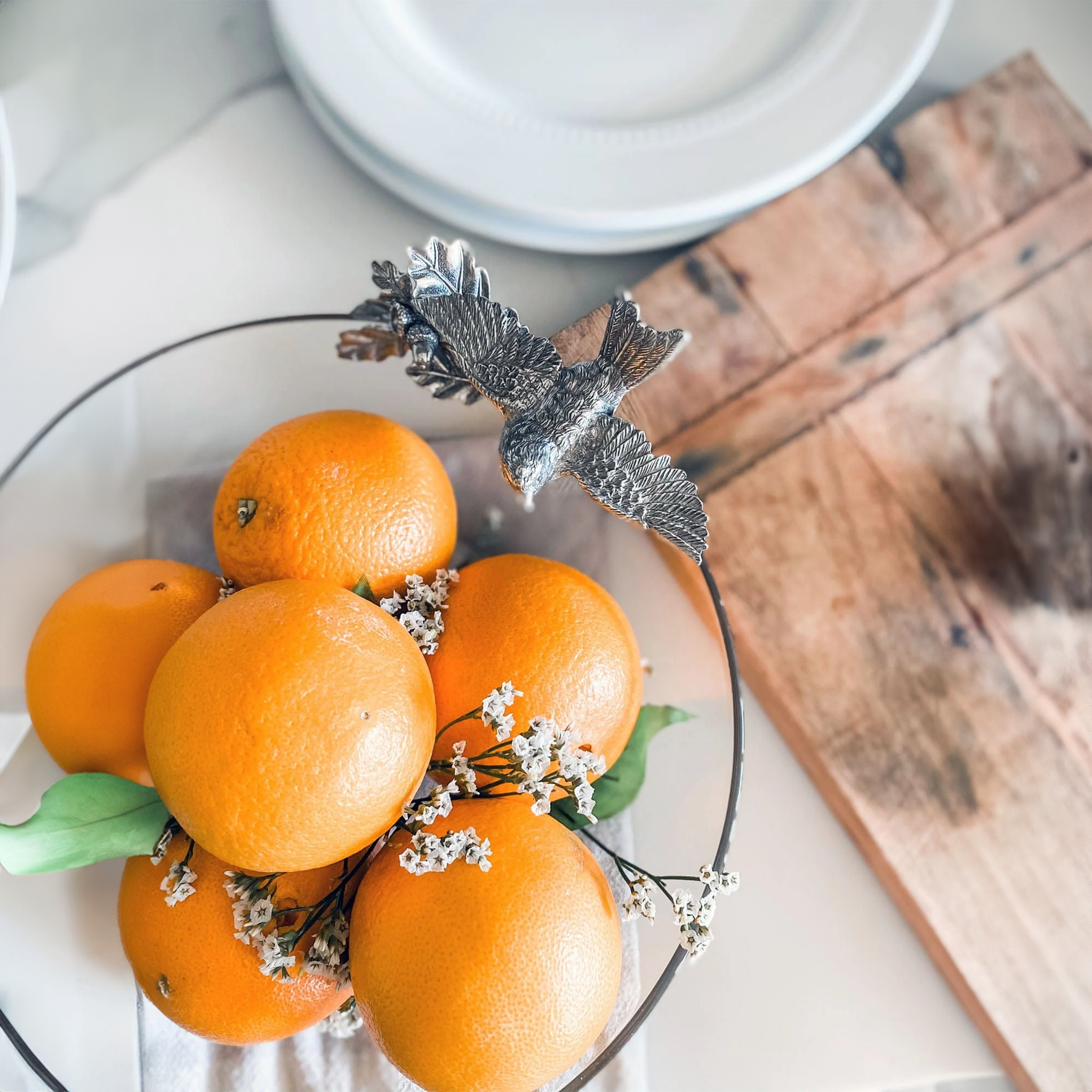 a glass serving bowl with a pewter bird on it filled with oranges on top of a table